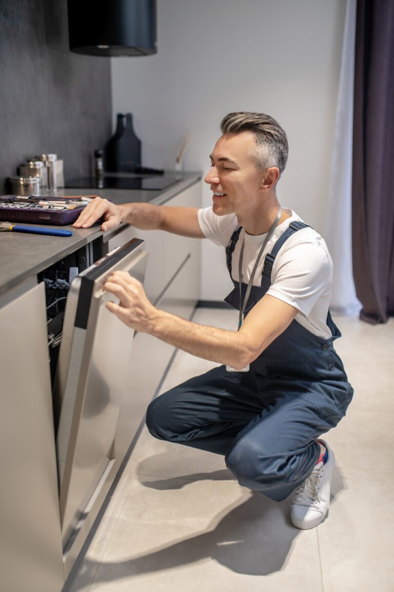 Working mood. Smiling man in tshirt and overalls crouching peeking into open door of built-in dishwasher in modern kitchen